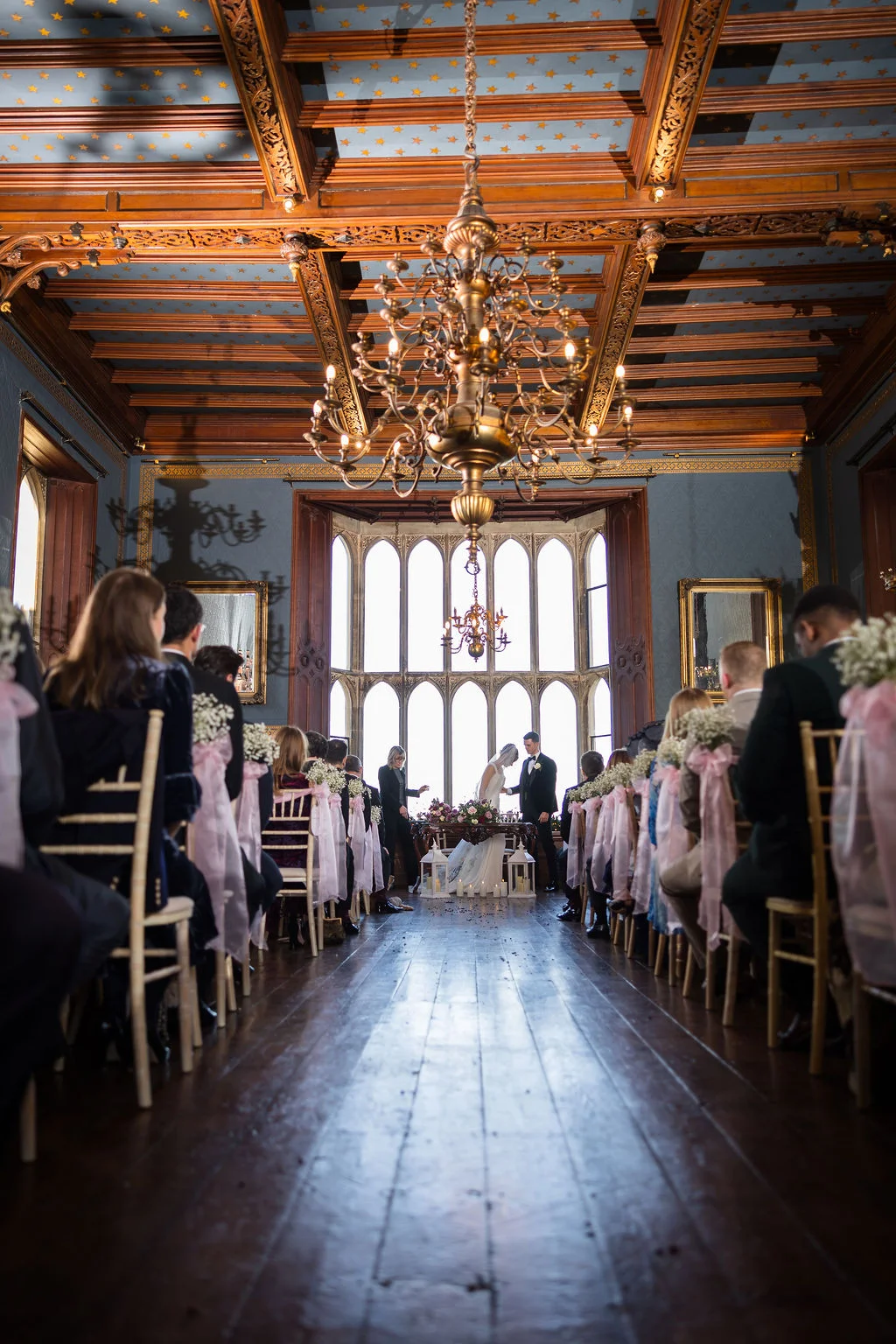 A wide angle view of an indoor wedding ceremony taking place in a grand panelled room with a starry blue ceiling and large arched windows.