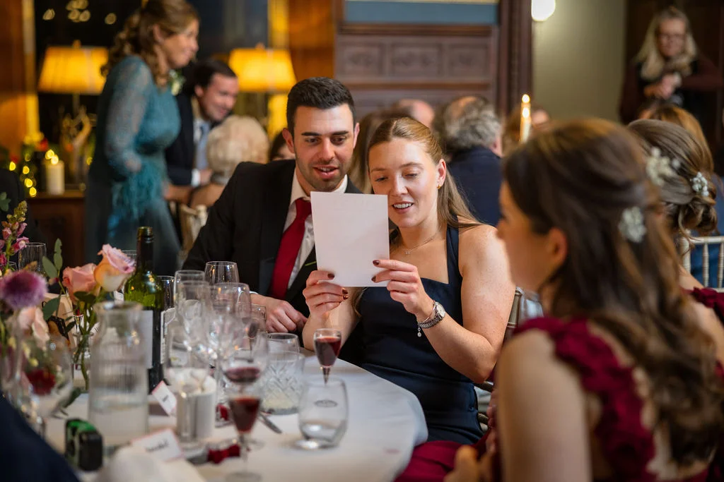 Wedding guests laughing together around a beautifully set dining table during speeches, illuminated by warm indoor lighting.