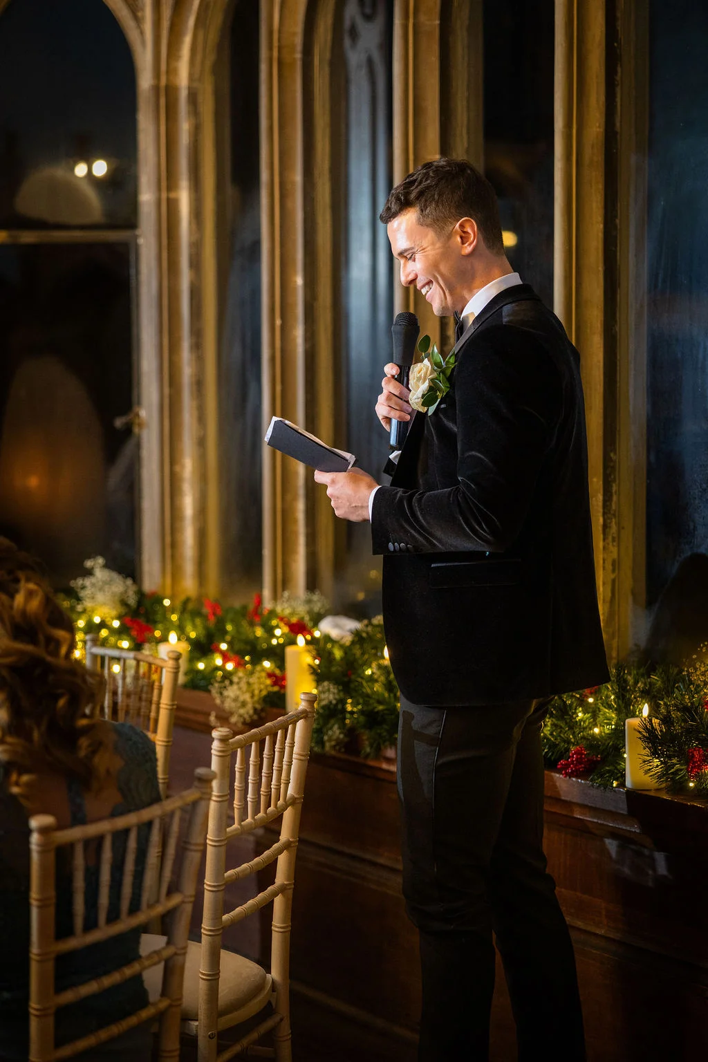 A smiling groom delivers his wedding speech, holding a microphone and notes while standing in front of tall arched windows adorned with festive greenery and candles.
