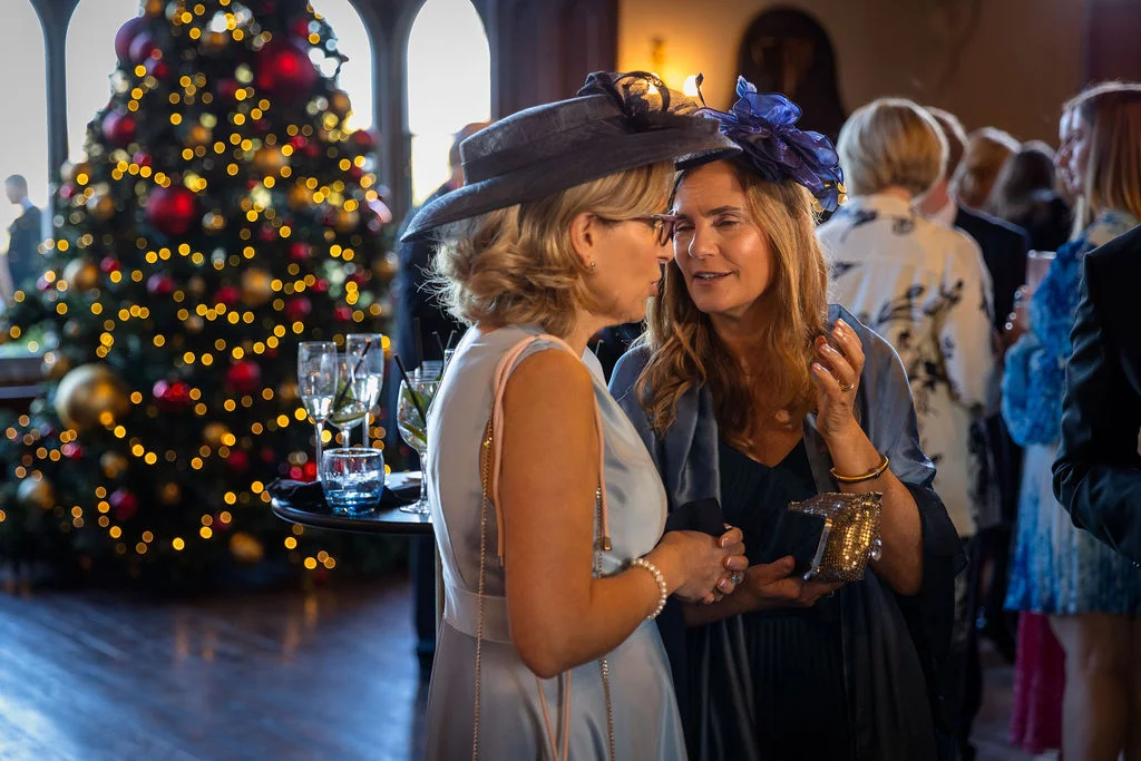 Two wedding guests wearing elegant dresses and fascinators chat near a glowing christmas tree during a festive winter drinks reception.