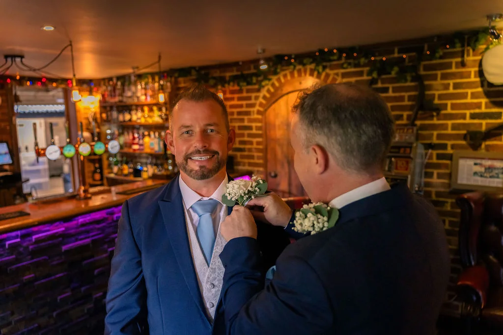A smiling groom in a blue suit and waistcoat has his buttonhole adjusted by another man inside a cosy pub wedding venue.