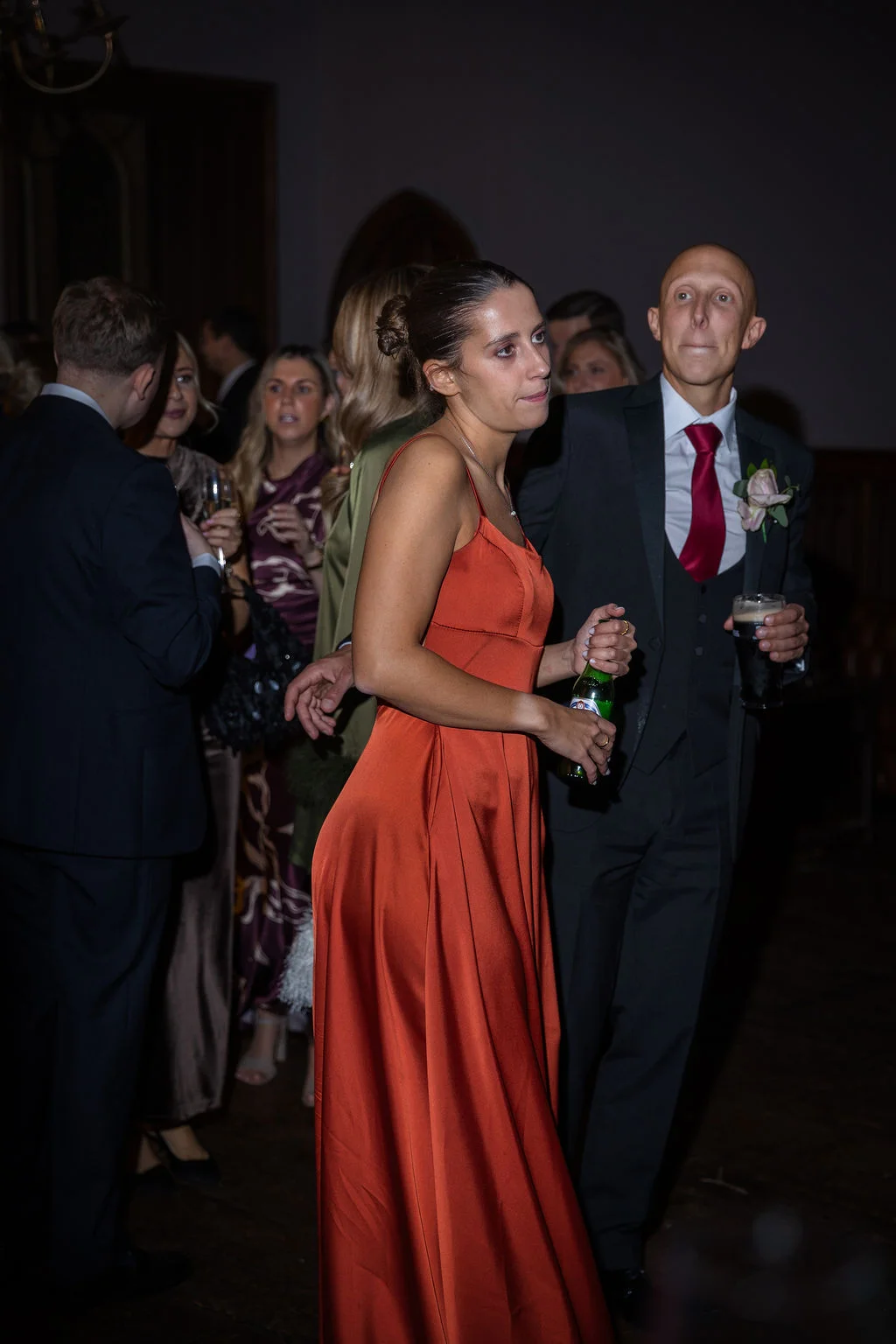 Guests mingling and holding drinks during an evening wedding reception, with a woman in a burnt orange satin dress in the foreground.
