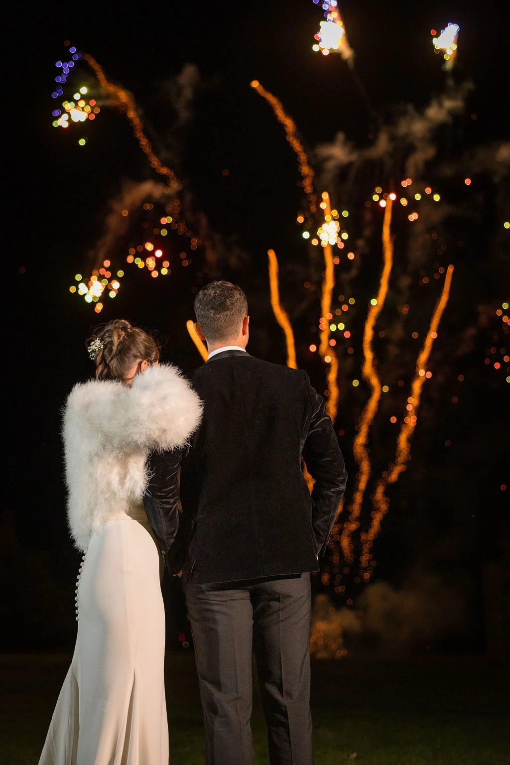 A newlywed couple stands back to back, watching a vibrant fireworks display light up the night sky during their winter wedding reception.