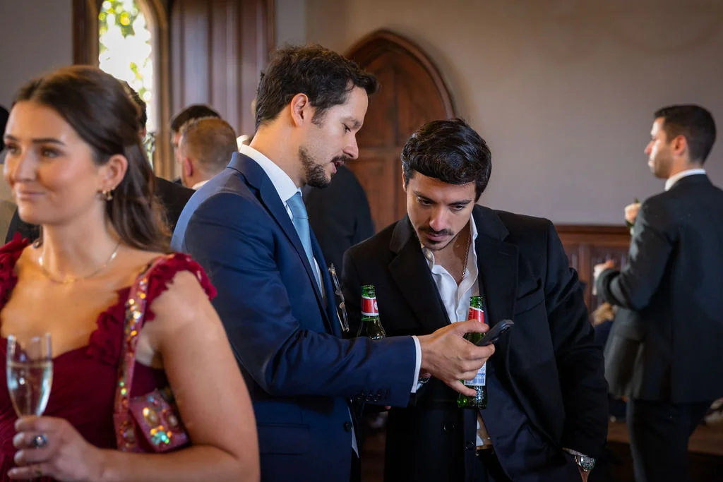 Wedding guests mingling indoors while two men in suits look down at a smartphone together, holding green glass bottles.