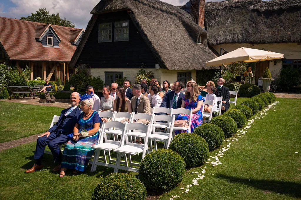 Wedding guests are seated on white folding chairs on a manicured lawn lined with topiary bushes and petal scatter for an outdoor ceremony.