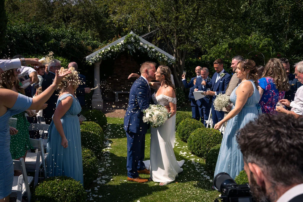 Newlyweds share a kiss at the end of an outdoor ceremony as family and friends shower them with white confetti in a sunlit garden.