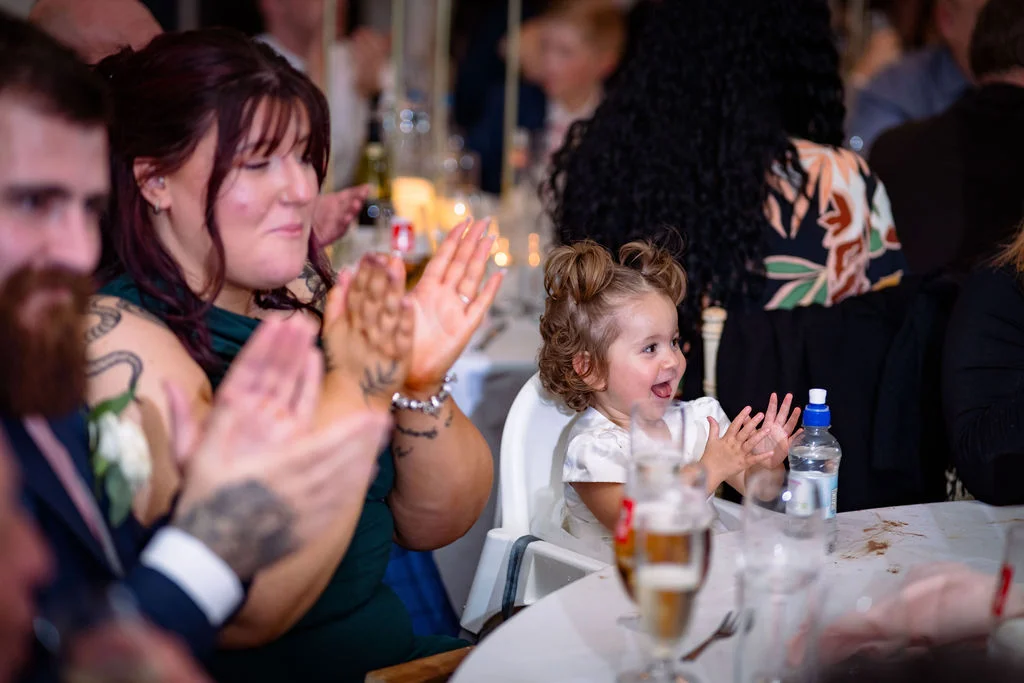 A young toddler sits in a high chair at a wedding reception, clapping and smiling enthusiastically alongside guests during the speeches.