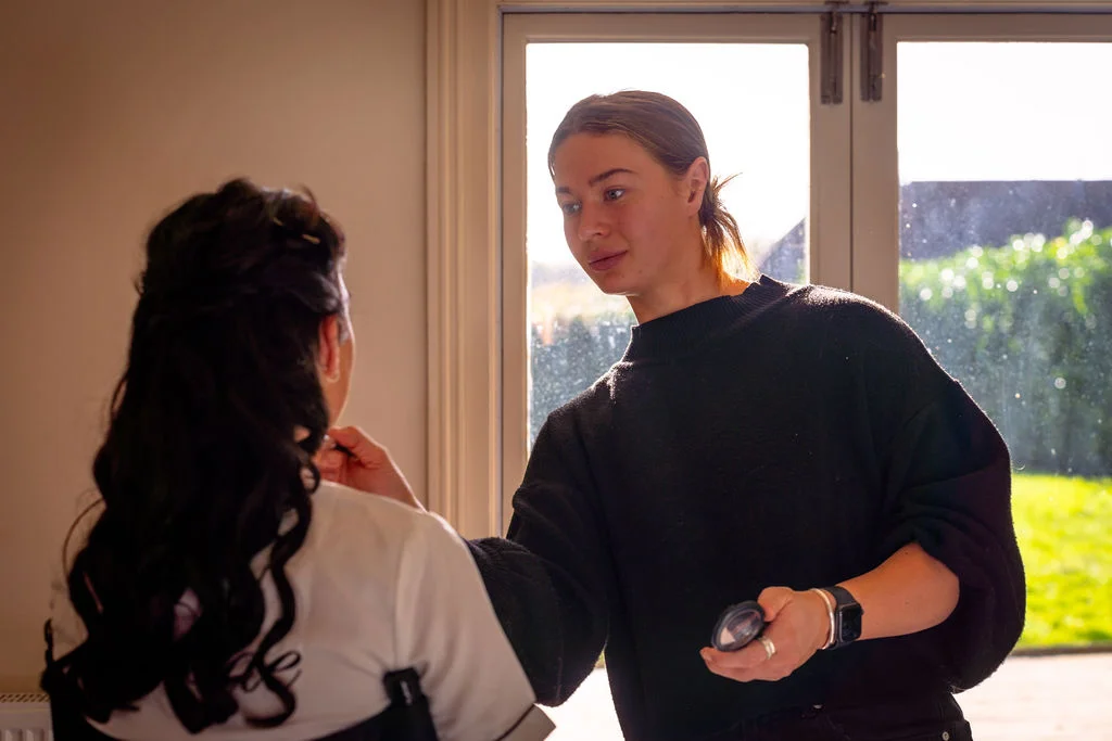 A professional makeup artist applies finishing touches to a bride sitting near a bright window during bridal preparations.