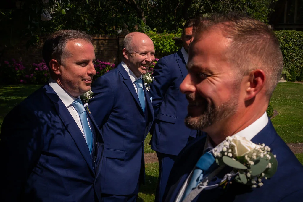 A group of smiling groomsmen wearing matching navy suits and blue ties adorned with white rose buttonholes stand outdoors in the sunlight.