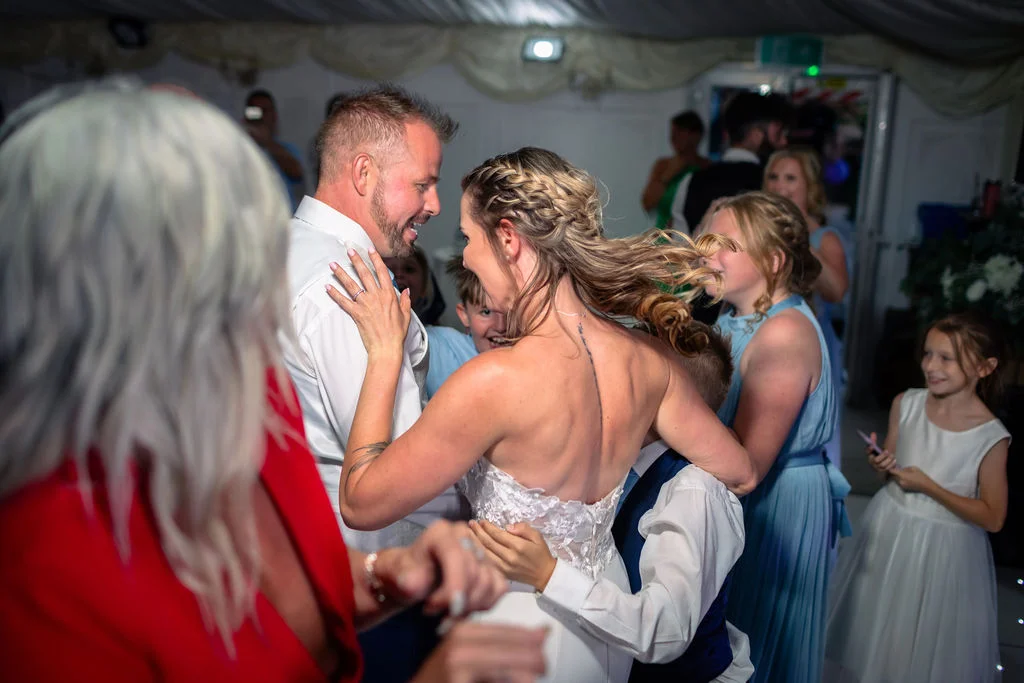 The bride and groom share a joyful moment dancing together at their evening wedding reception surrounded by family and guests.