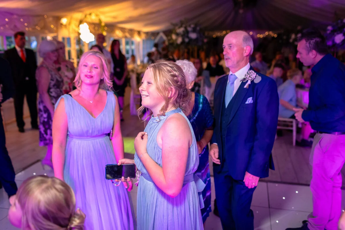 Wedding guests in blue attire and evening suits mingle and smile together inside a brightly lit marquee during the evening reception.