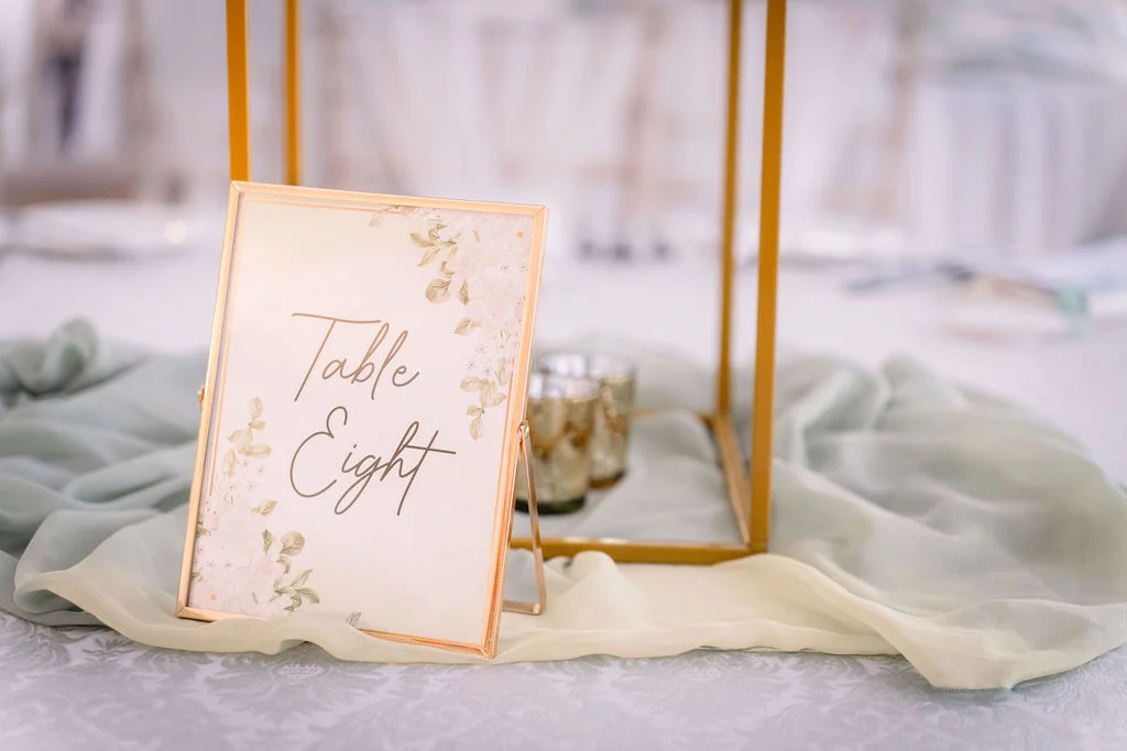 An elegant table number card reading Table Eight rests on a pale sage green runner alongside glowing votive candles and a modern gold geometric centerpiece frame.