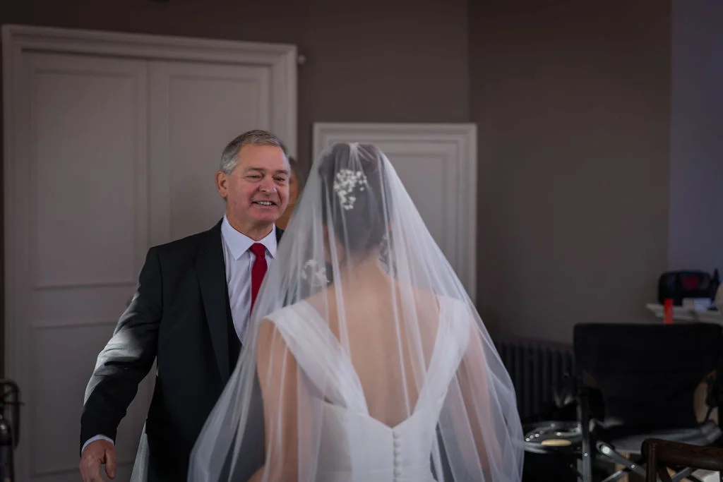 A smiling father sees his daughter in her wedding dress and veil for the first time during a touching bridal prep moment.