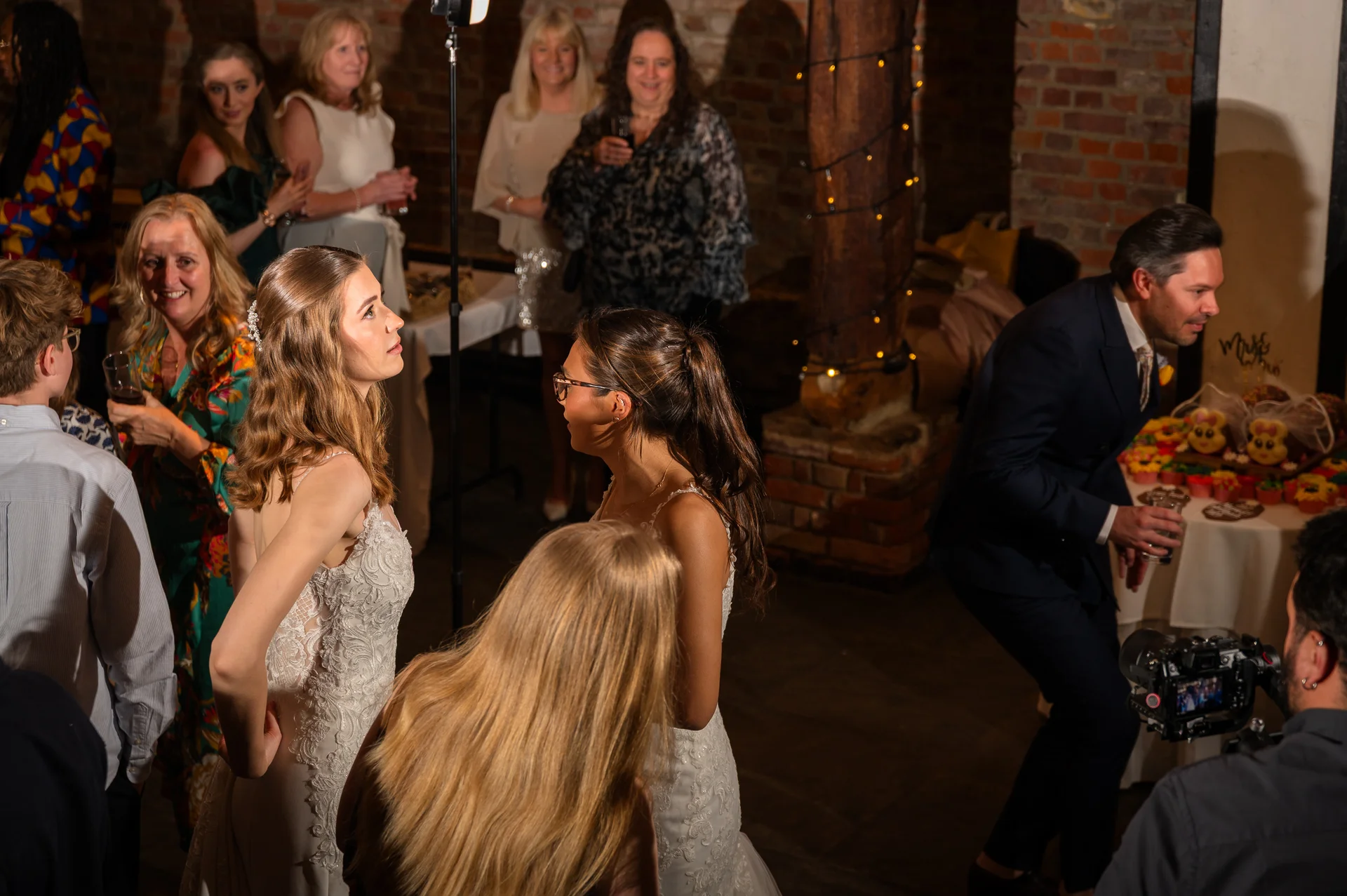Two brides in lace wedding dresses stand together surrounded by wedding guests in a rustic stone barn reception venue with exposed brick walls.