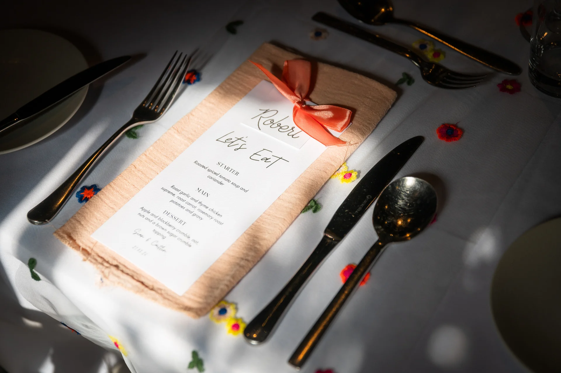 A beautifully set wedding place setting featuring a printed menu on a peach napkin tied with a satin bow, flanked by cutlery on a white cloth.