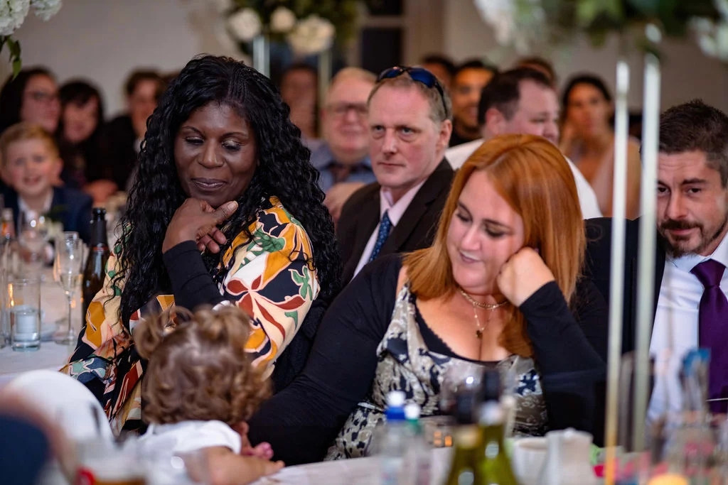 Wedding guests attentively watching the speeches during a reception dinner in a brightly lit venue.