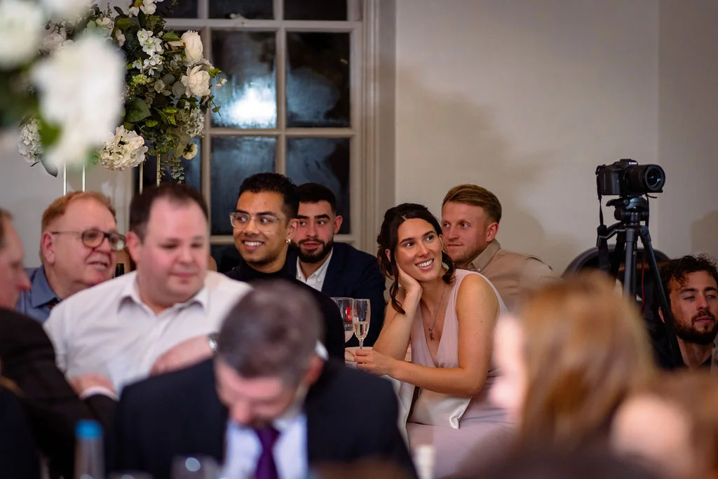 Wedding guests seated at tables smiling and watching speeches unfold during the wedding breakfast reception.