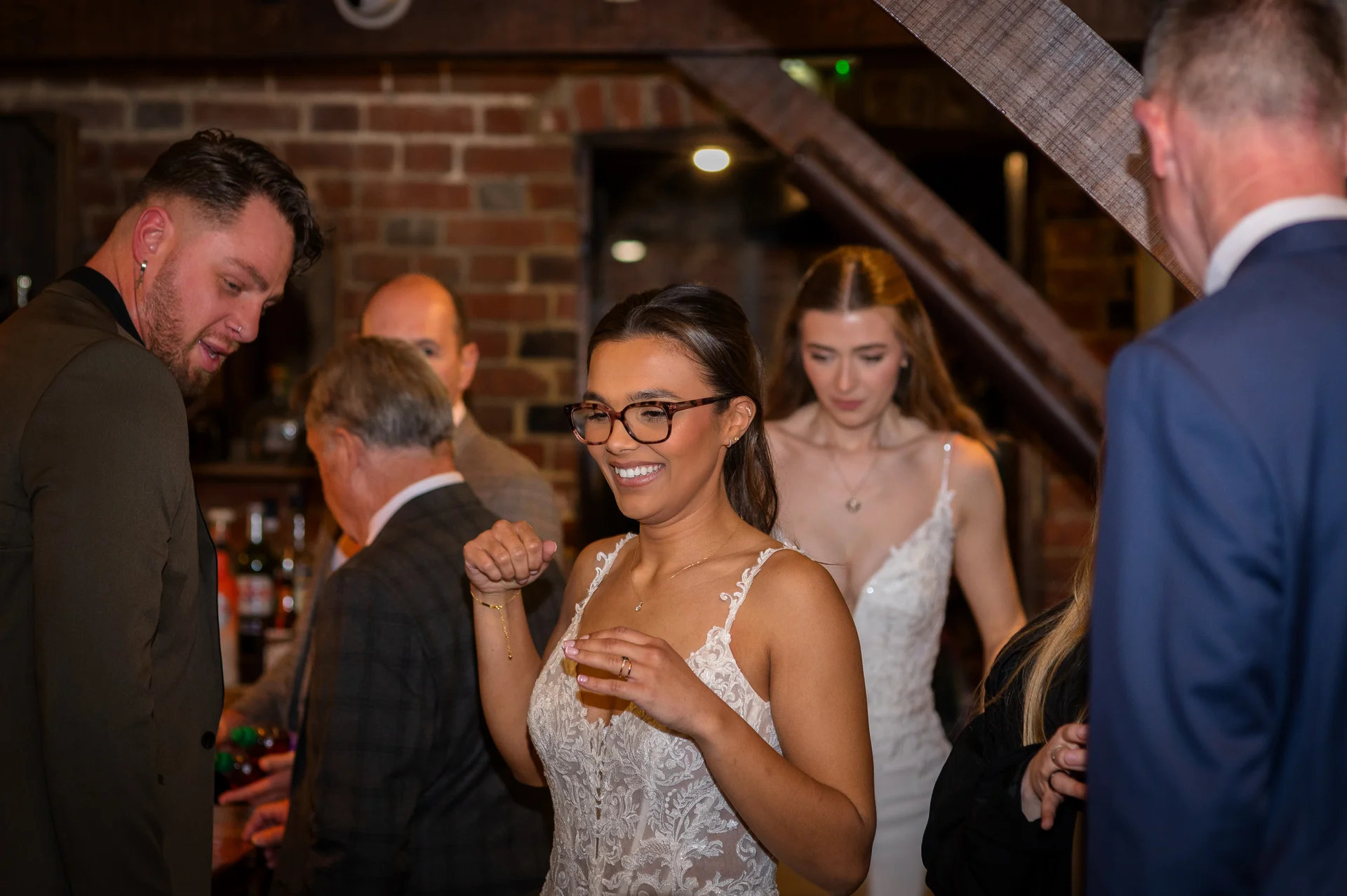 A smiling guest wearing glasses and a lace gown laughs while mingling with friends inside a rustic barn venue featuring exposed brick walls.