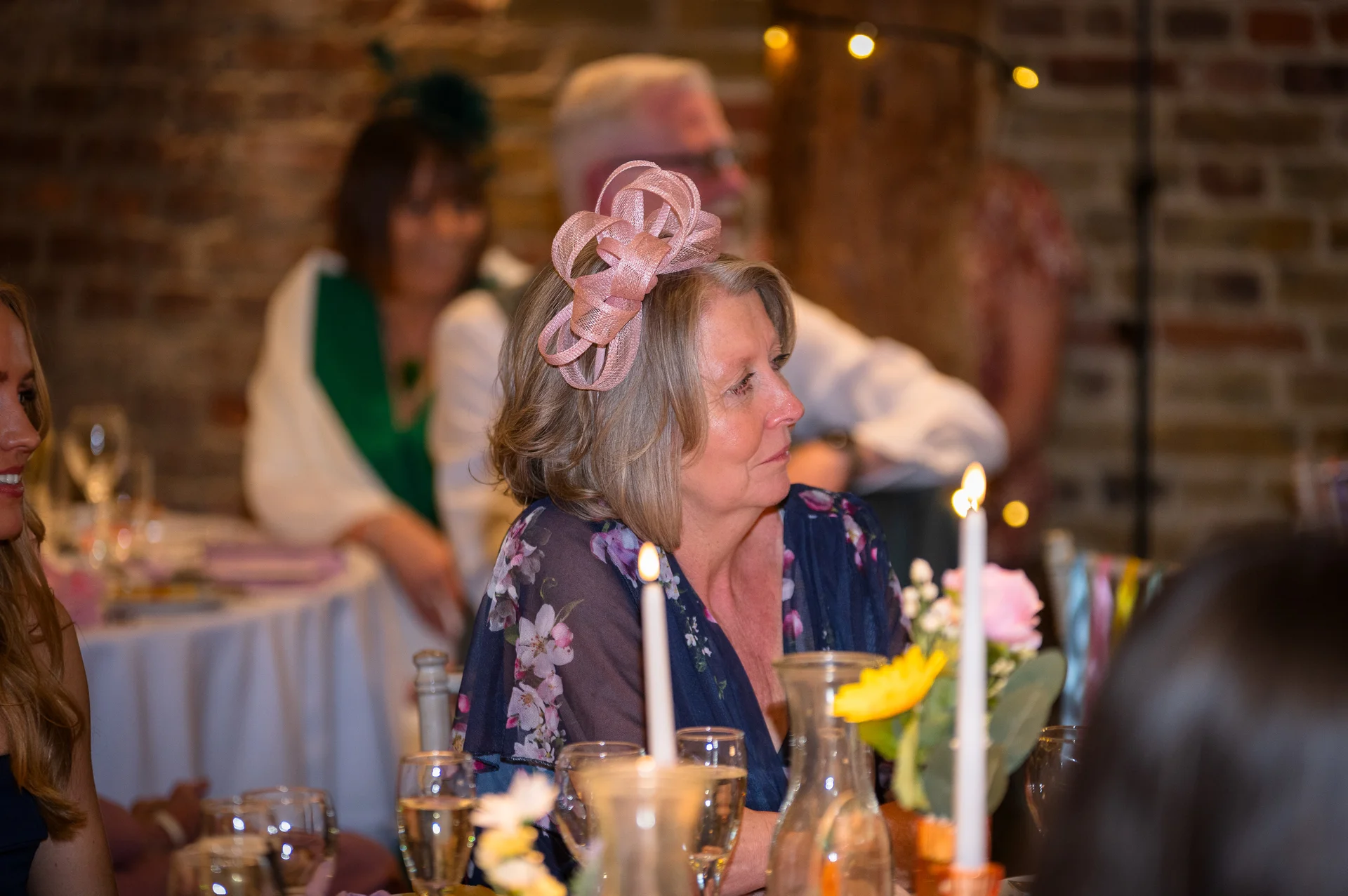 A guest wearing a pink fascinator and a floral dress sits attentively at a candlelit wedding breakfast table against an exposed brick background.