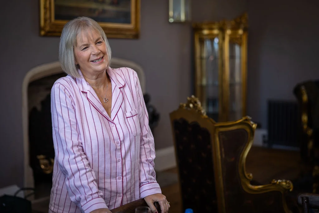 A mature woman with short grey hair smiles happily while wearing pink striped pyjamas in an elegantly decorated indoor venue during morning preparations.