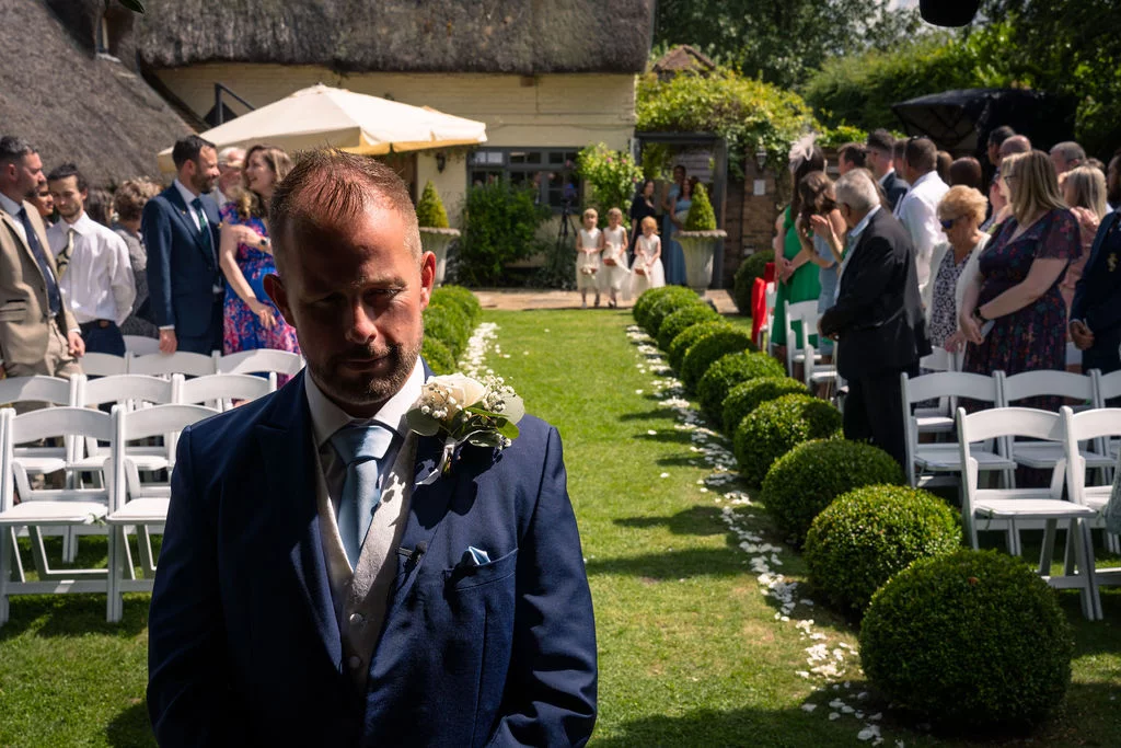A groom in a navy suit and waistcoat waits anxiously at the top of an outdoor aisle lined with white chairs and neatly trimmed box balls.