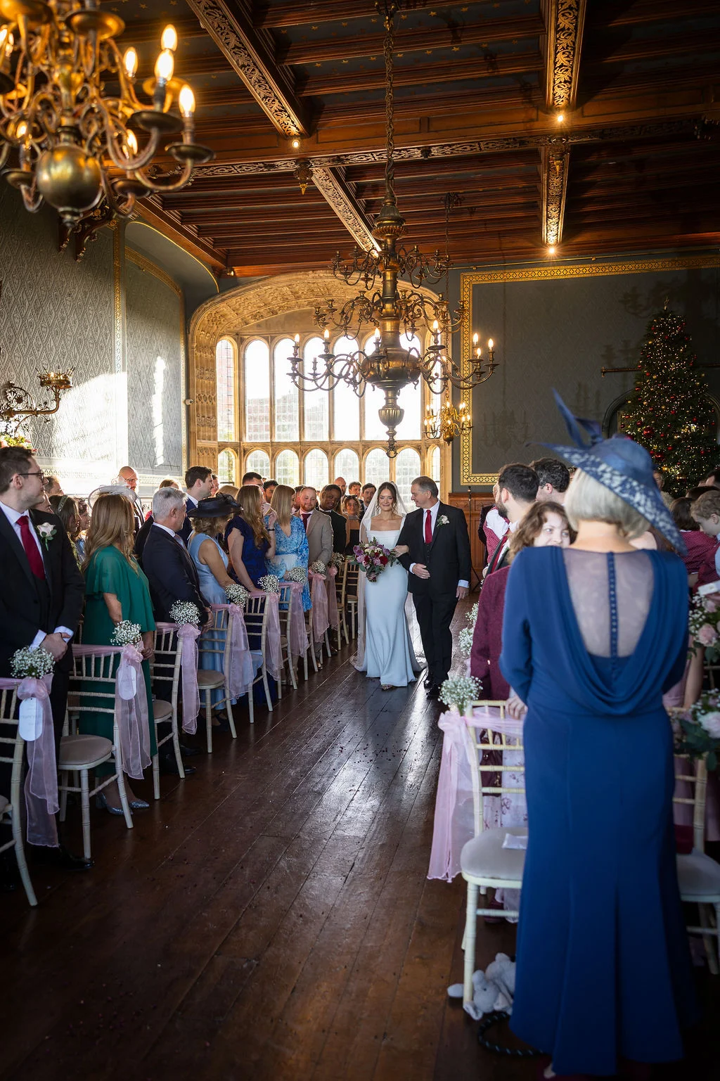 The bride walks down the aisle accompanied by her father inside a grand historic hall featuring a high wooden vaulted ceiling, magnificent chandeliers, and guests seated on either side.