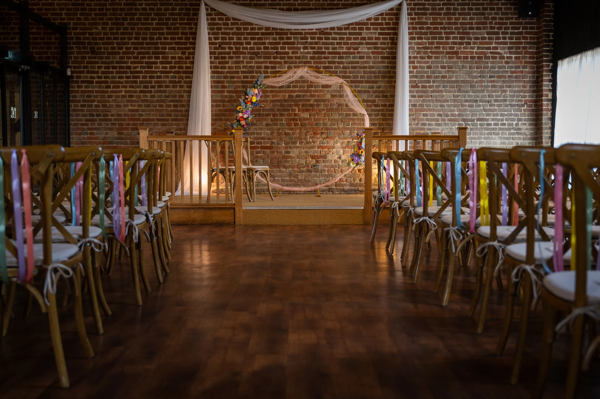An empty wedding ceremony setup featuring wooden cross-back chairs decorated with colourful pastel ribbons lining an aisle towards a circular floral arch against an exposed brick wall.