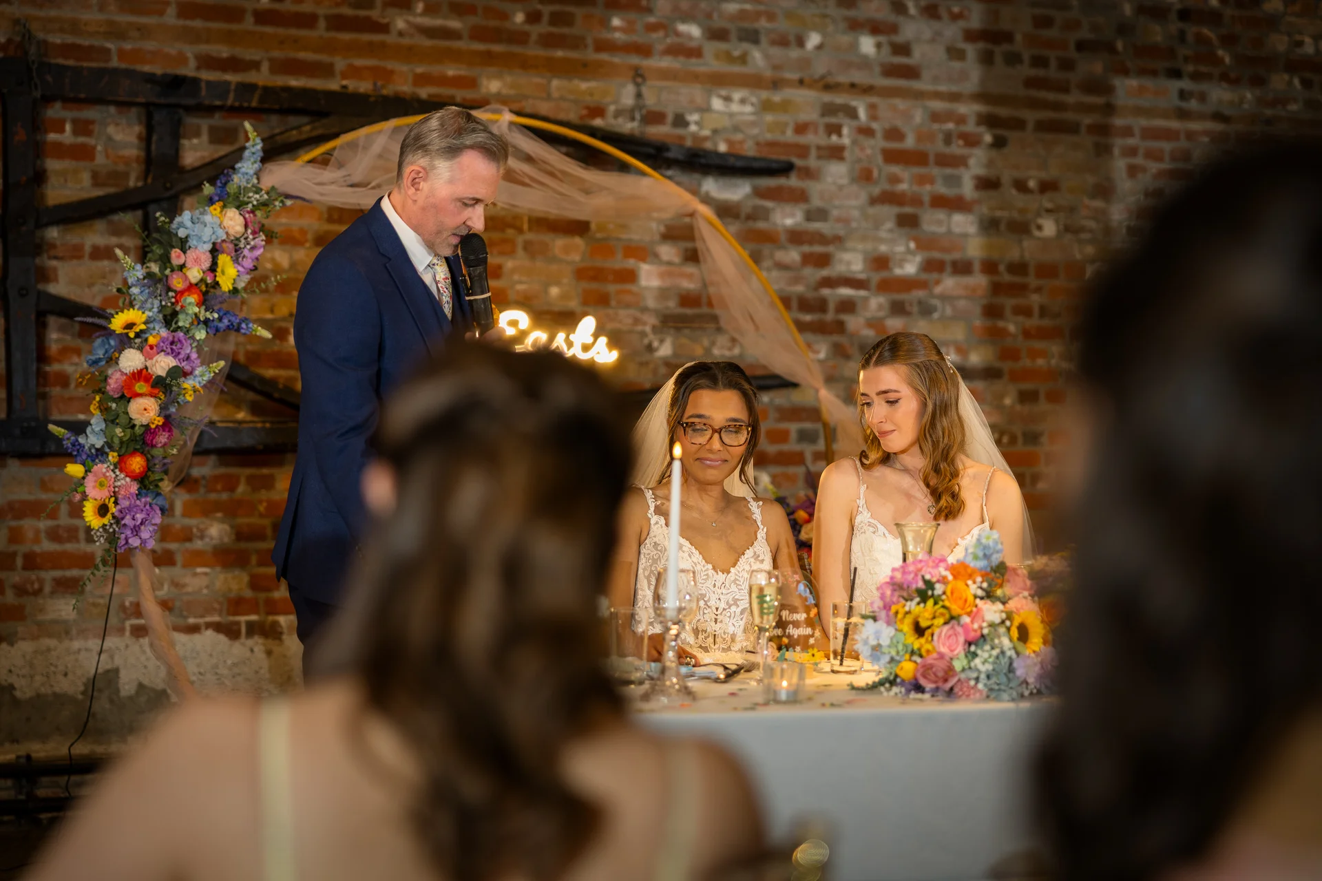 An emotional father gives a speech while two brides sit together at the top table during their wedding reception in a rustic barn venue with exposed brick walls.