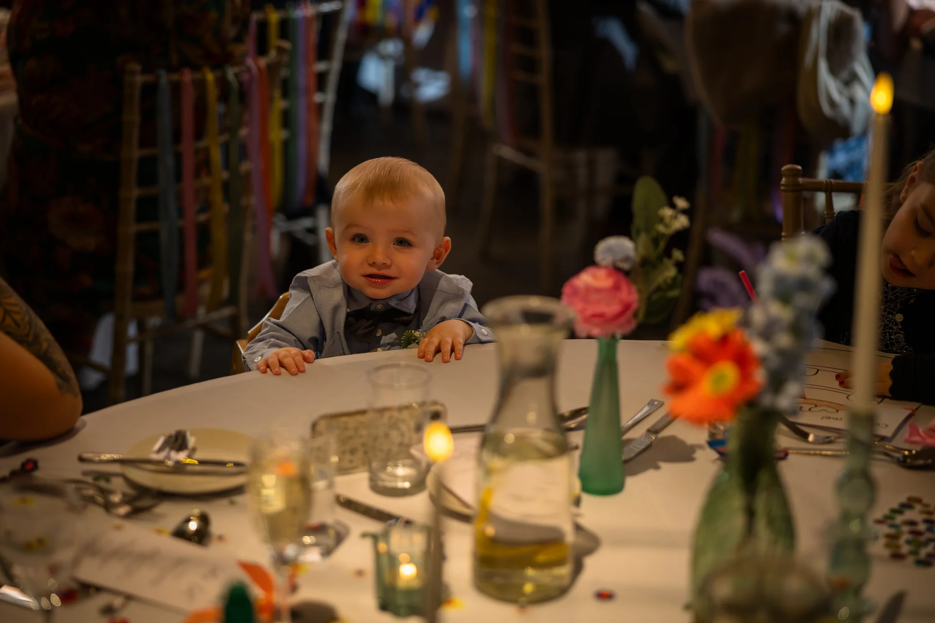 A young page boy with fair hair sits at a decorated round wedding table, looking curiously towards the camera amidst colourful floral bud vases and glassware.