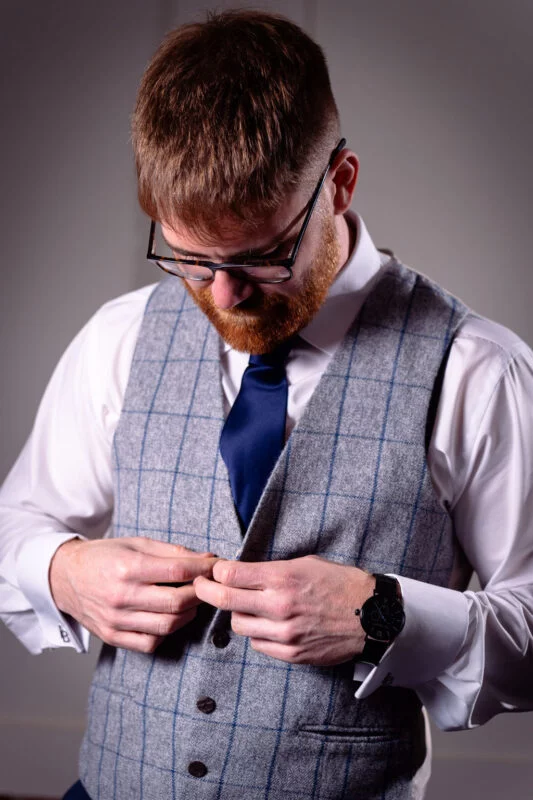 A groom with a red beard and glasses carefully buttons his grey tweed waistcoat while wearing a crisp white shirt and a navy tie during morning preparations.
