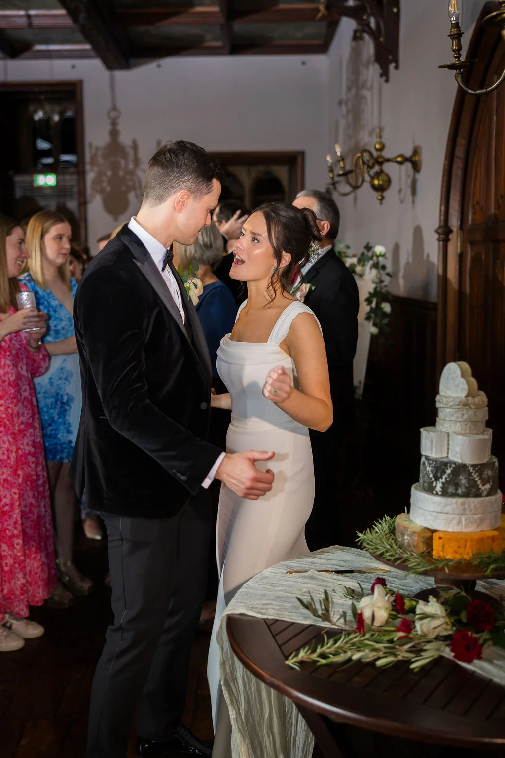 A bride and groom react joyfully while standing near a tiered cheese wedding cake during an evening reception in a historic country house.