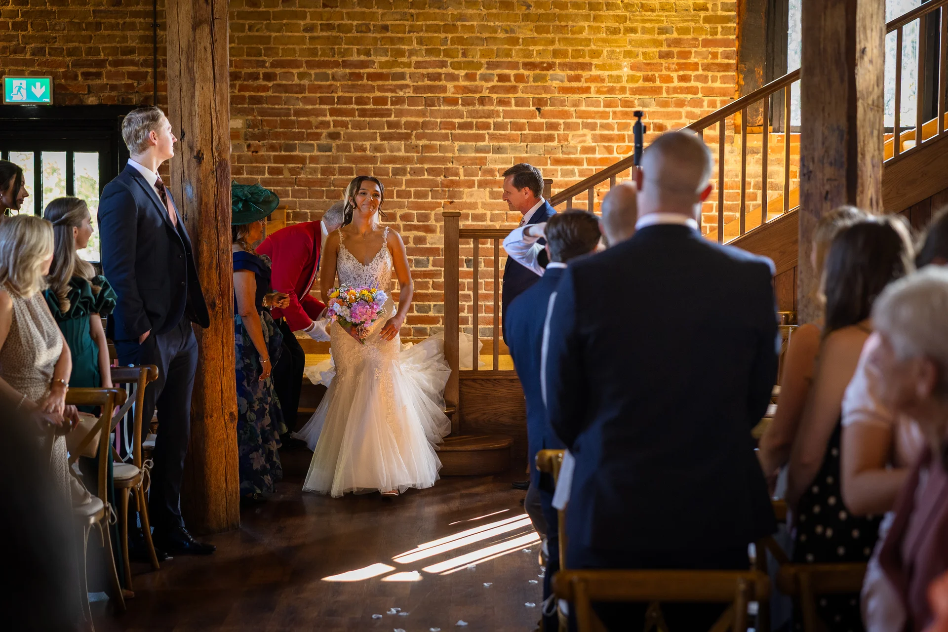 A smiling bride holding a colourful bouquet walks down the aisle in a rustic brick barn venue towards her waiting groom.