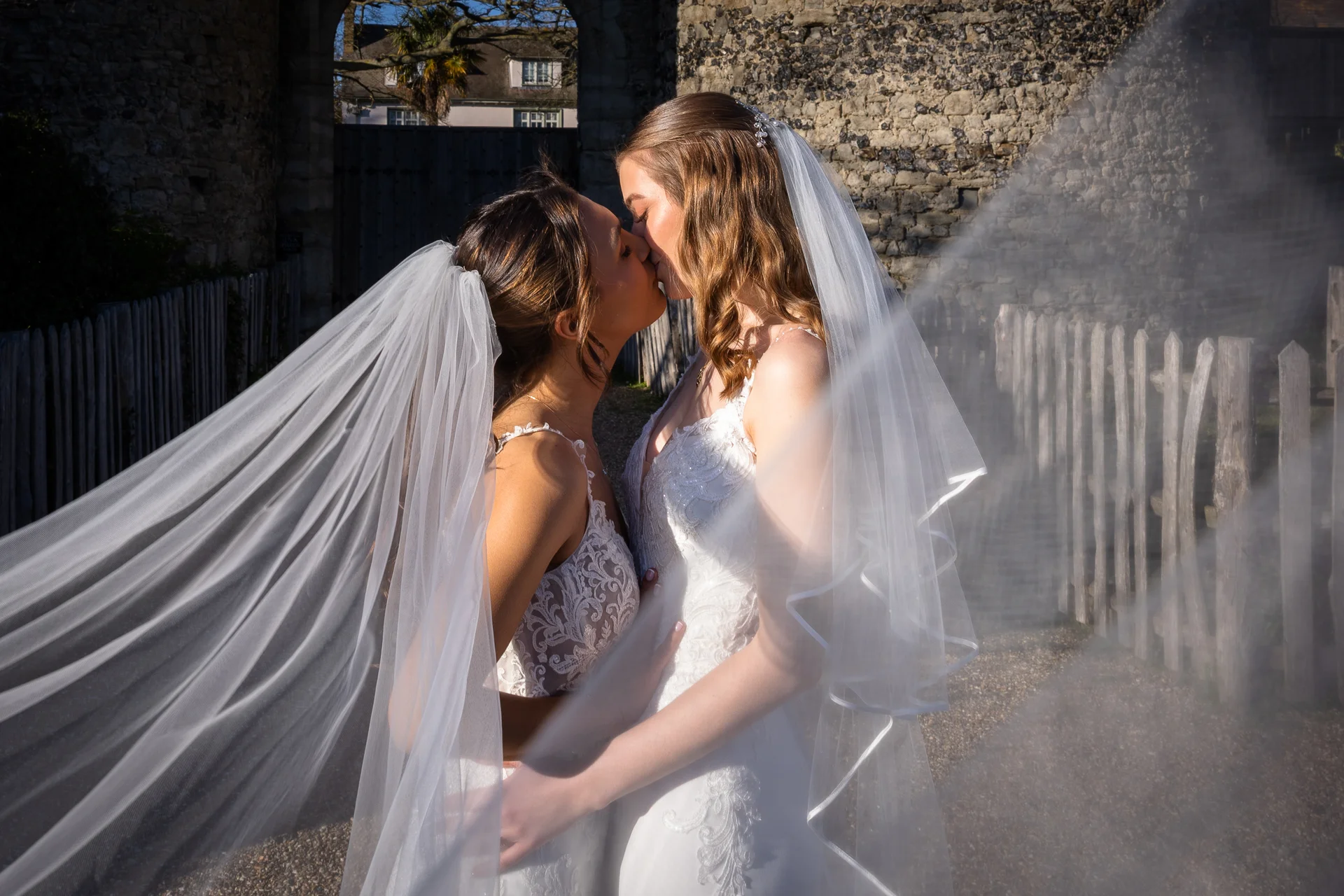 Two brides share a tender kiss outdoors in front of a stone archway, framed artistically by a sheer veil caught in the wind.