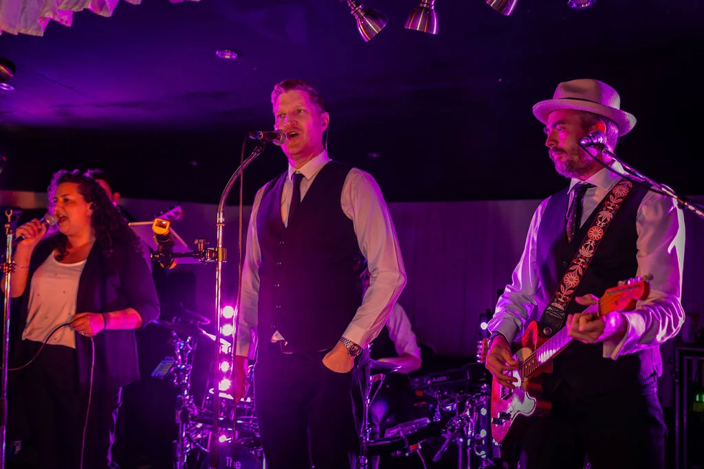 A live wedding band performs on stage under dramatic purple lighting, featuring a male vocalist and a guitarist in waistcoats.