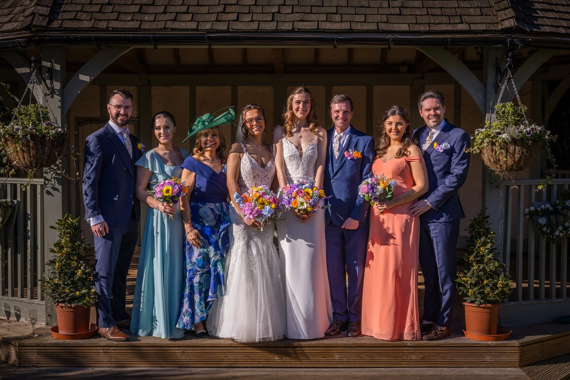 A full-length group portrait of two brides, their wedding party, and parents standing on a wooden deck in front of a gazebo on a sunny day.