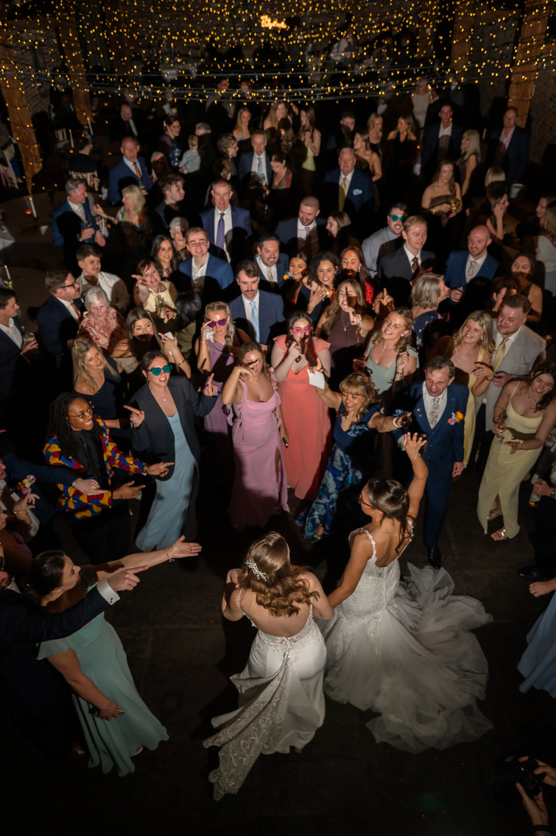 An aerial view of two brides dancing surrounded by cheering guests under a dazzling canopy of fairy lights.