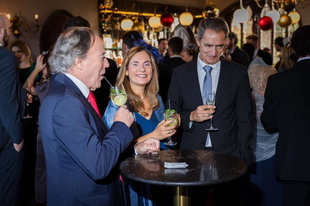 Wedding guests wearing formal attire and smiling while enjoying cocktails and mingling at an indoor evening reception.