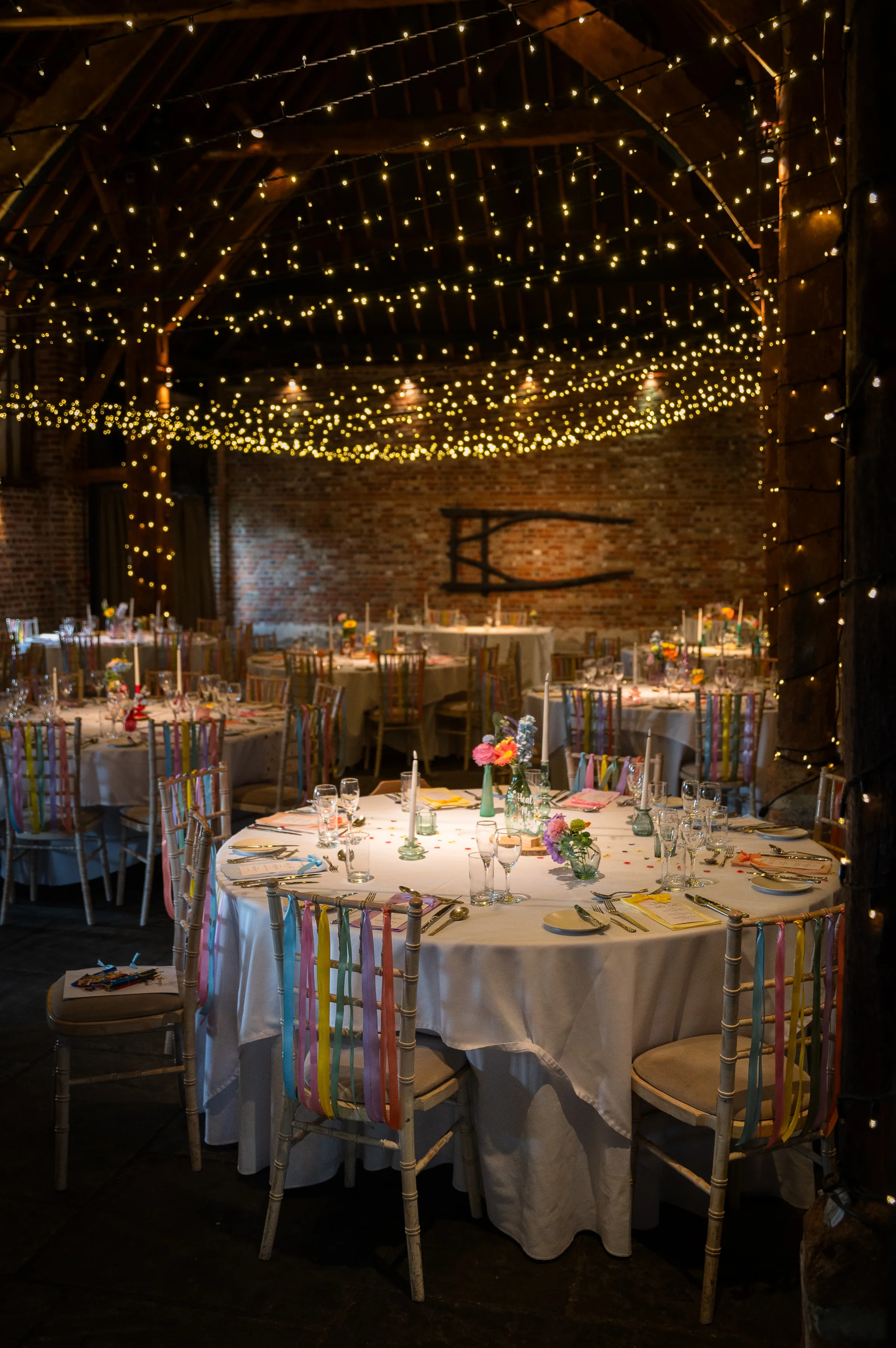 A warmly lit rustic barn wedding reception features round dining tables adorned with white linens, colourful ribbon-decorated chairs, and a dazzling canopy of fairy lights overhead.