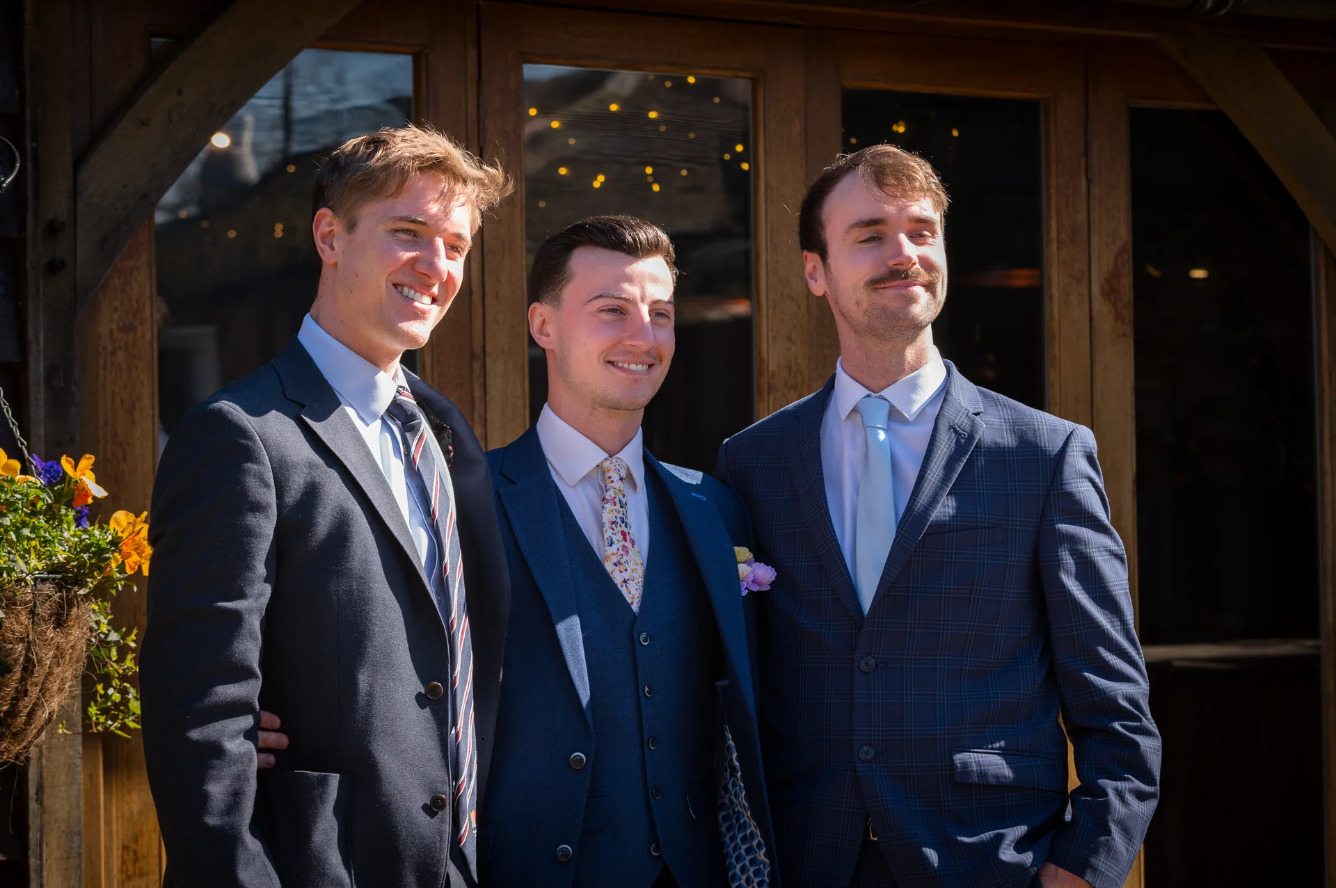 Three smiling groomsmen stand together outside a rustic wooden barn venue in matching tailored suits under bright sunshine.