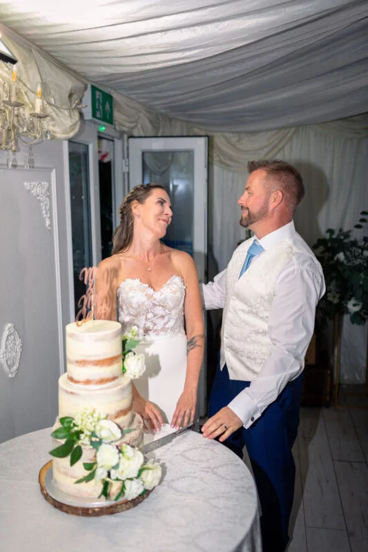 The newly married couple lovingly look at each other while cutting a tiered semi-naked wedding cake adorned with white florals inside a marquee.