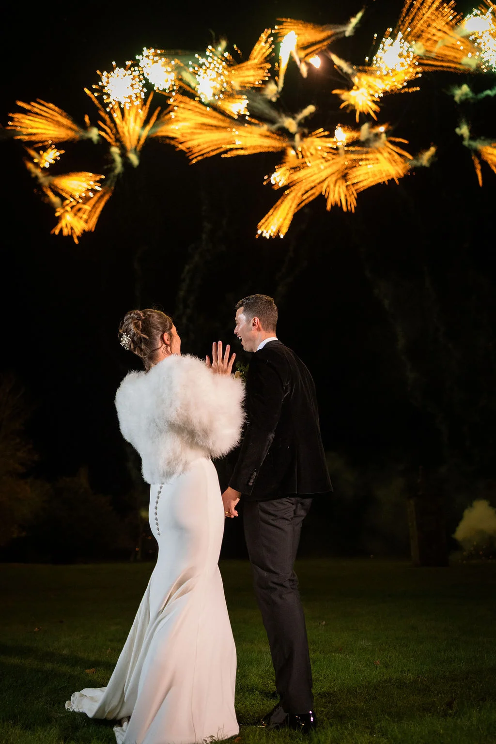 A newlywed couple stands outdoors at night admiring a spectacular fireworks display exploding across the dark sky above them.