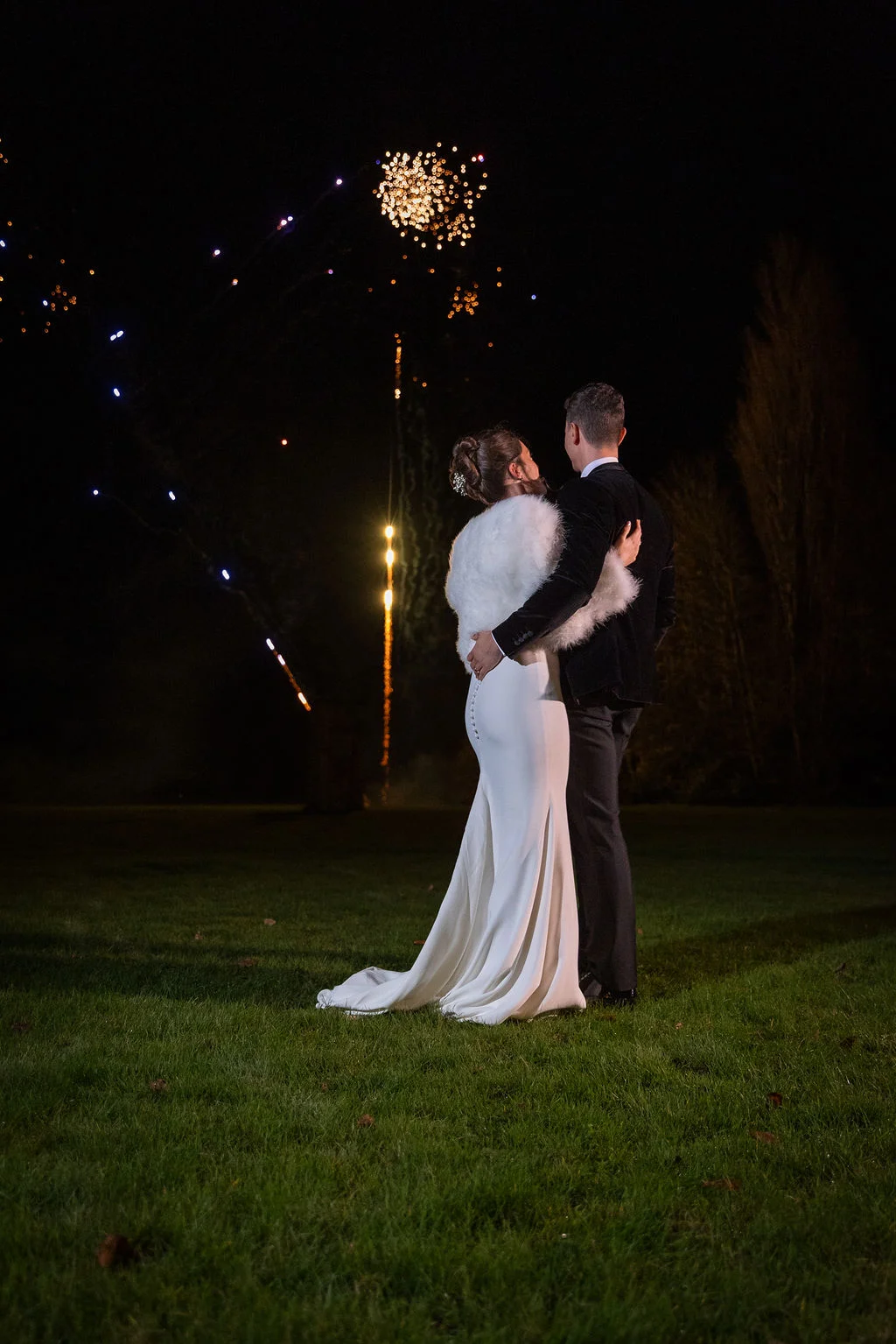 A newlywed couple embraces on a lawn at night, watching a vibrant fireworks display light up the dark sky during their winter wedding.