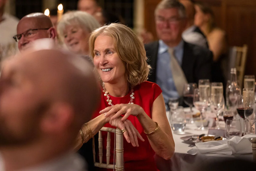 A smiling guest wearing a red dress and pearl necklace enjoys the wedding speeches at a banquet table.