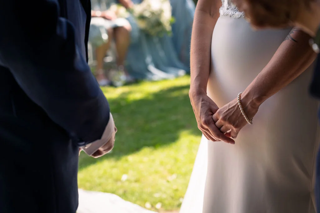 A close-up shot captures the tender moment of a bride and groom standing together during an outdoor ceremony, with the bride wearing a sleek crepe gown and a delicate pearl bracelet.