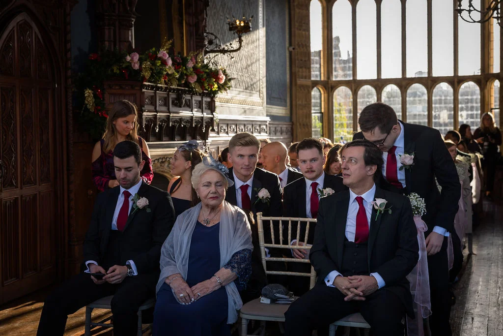 Family members and guests are seated in a grand, wood-panelled room with large arched windows during a wedding ceremony.