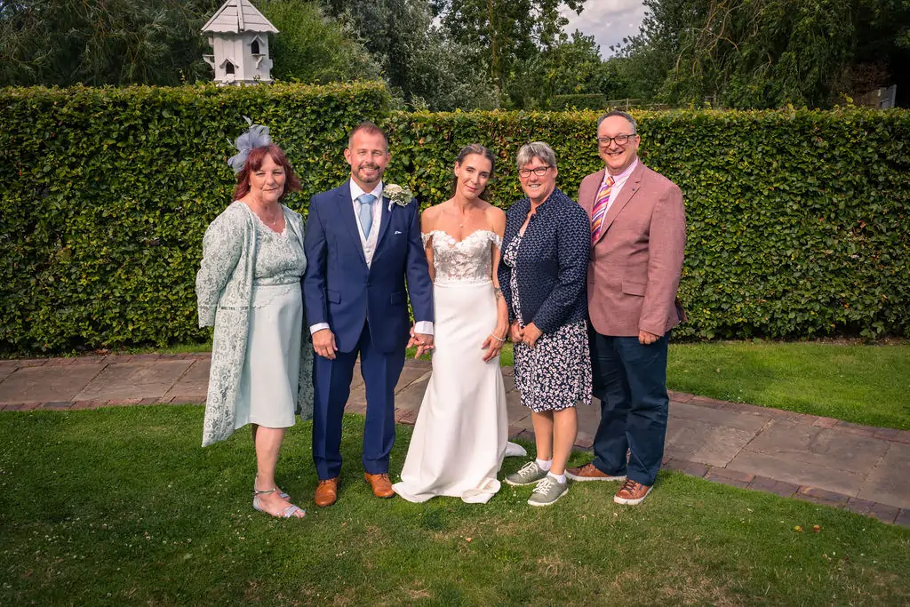 A newlywed couple poses outdoors in a manicured garden with family members against a tall green hedge backdrop on their wedding day.