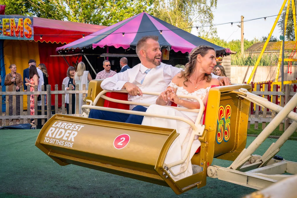 A bride and groom laugh together while riding a vintage fairground attraction at a festival-themed wedding.