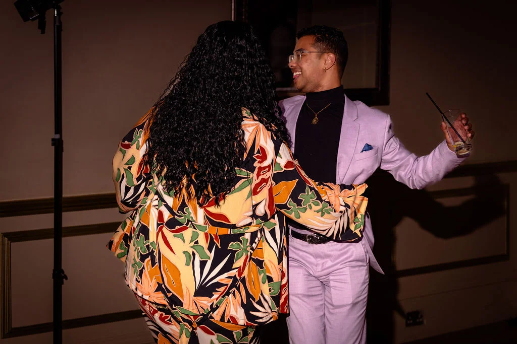 A guest in a bold floral suit dances happily with someone wearing a light purple suit and black turtleneck while holding a drink at an evening reception.