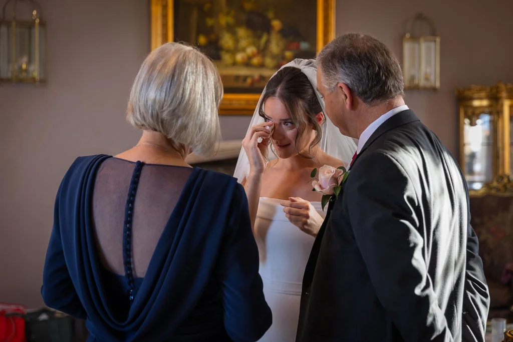 An emotional bride gently wipes away tears as she shares a tender moment with her parents during wedding preparations indoors.