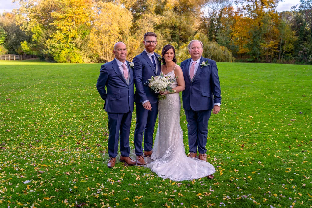 A newlywed bride holding a bouquet stands on a green lawn scattered with autumn leaves alongside her groom and two family members in matching navy suits.